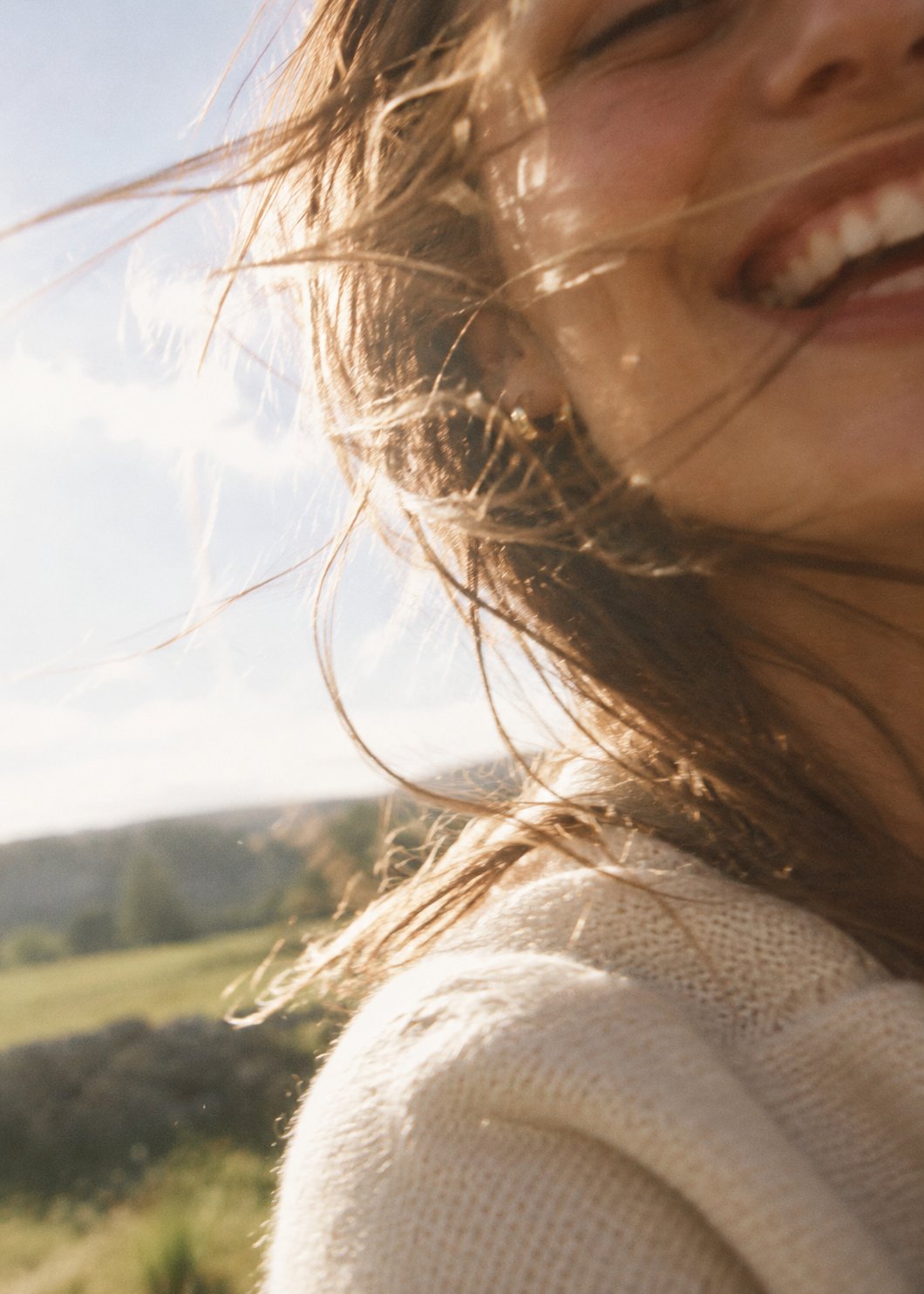 A smiling woman with hair blowing across her face in the wind, sunlit field behind her