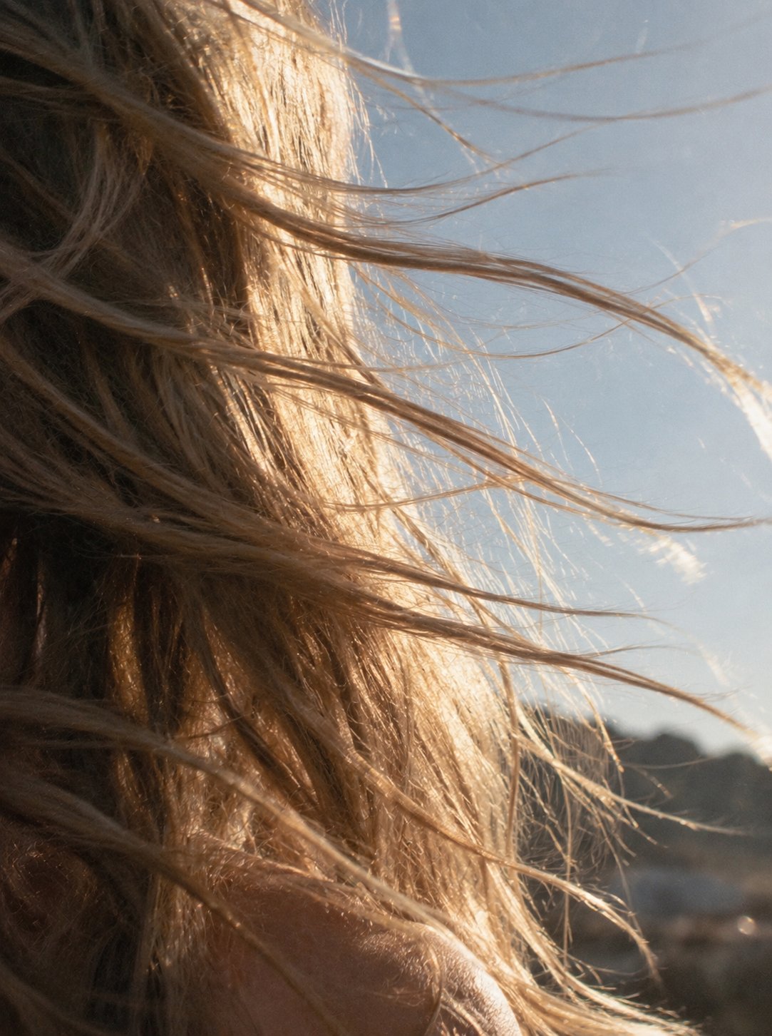 Blonde hair blowing in the wind, backlit by warm sun against a pale sky